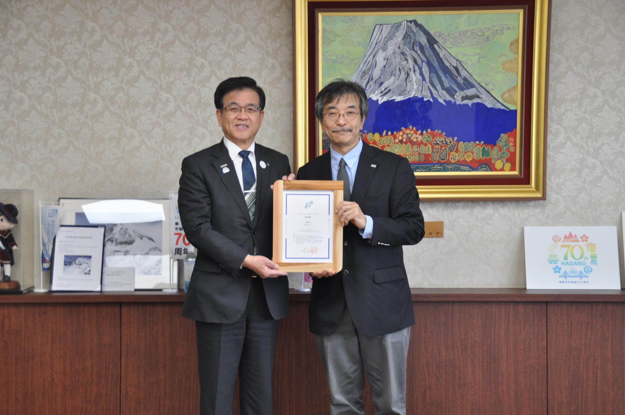 山の絵画が掛かった壁を背景に、スーツ姿の市長と男性が、一緒に木製の額縁に入った認証書を持って並んで立っている記念写真