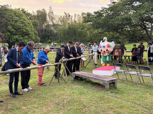 大きな木々や竹林に囲まれた芝生広場で、中井町イメージキャラクターのなかまるや複数の来場者が見守る中、一列に並んだ7人の男性たちが、横向きに置かれた竹灯籠に点火を行っている様子の写真