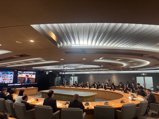 照明に特徴のある会議室でU字型の机を囲んで多くの関係者が座り、前方に2つ並んだ大きなモニターに、オンライン出席者たちと、発言中の市長のアップが映し出されている会議の様子の写真