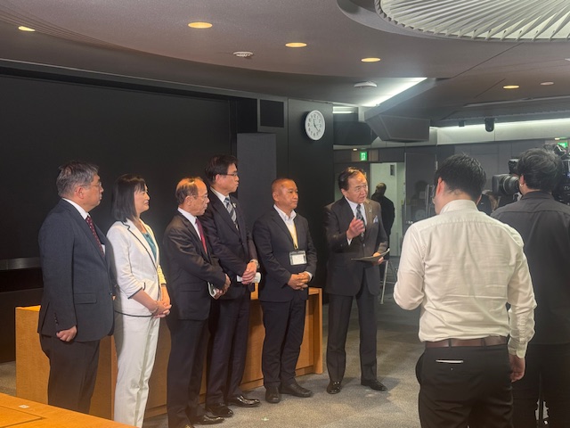 6人のスーツ姿の男女が横一列に並んでおり、向かって右端に立つ男性が撮影している報道陣のカメラに向かって話している様子の写真