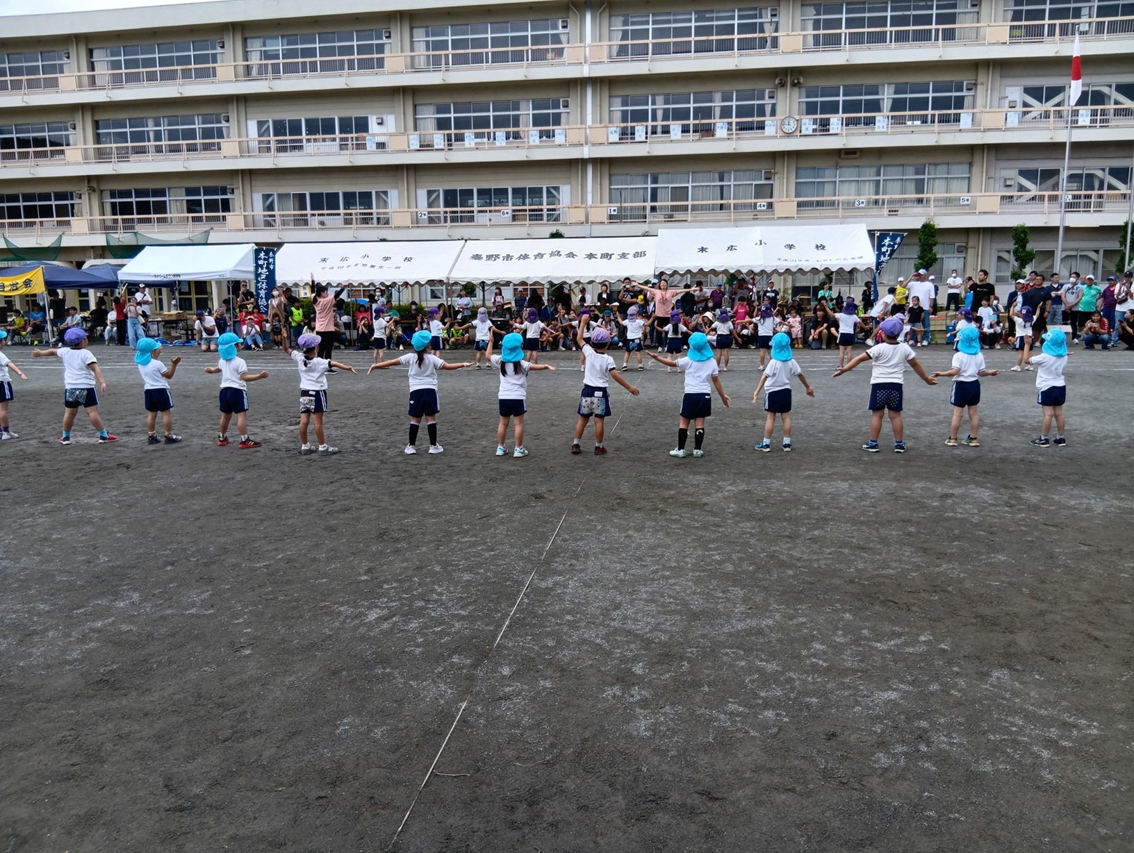 4階建ての白壁の校舎の前に、大勢の観客や関係者が集まった白いテントが並び、その前のグラウンドでは水色や紫色の帽子をかぶった子どもたちが横一列に並び両腕を広げて立っている後ろ姿を撮影した写真