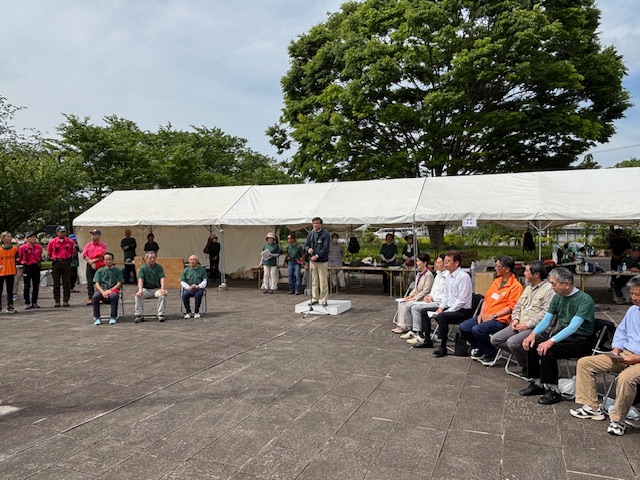 大きな木々に囲まれ、参加者や関係者が椅子に座ったり立っている白いテントが並んで設置された広場で、中央の白い台の前に設置されたマイクスタンドの前に立つ市長が挨拶をしている丹沢ボッカ駅伝競争大会開会式の様子の写真