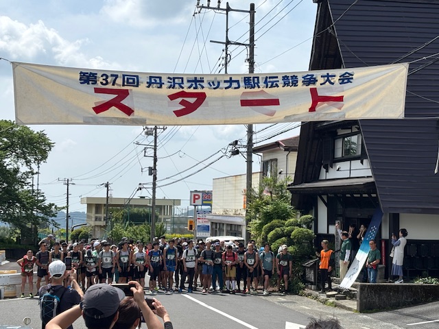 道路の上に「第37回 丹沢ボッカ駅伝競走大会 スタート」と書かれた横断幕が掲げられ、ゼッケンをつけた大勢のランナーがスタートラインに並んでいる、駅伝競争大会の様子の写真