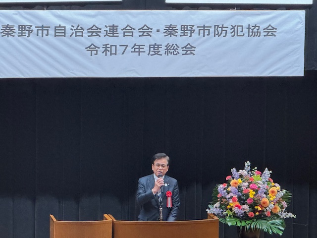 「秦野市自治会連合会・秦野市防犯協会 令和7年度総会」と書かれた横断幕が掲げられたステージの、色とりどりの花が飾られている演台で、マイクを持って挨拶するスーツ姿の市長の写真