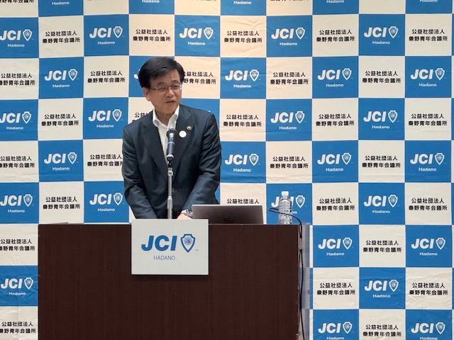 秦野青年会議所のバックパネルを背景に、演台の前に立つスーツ姿の市長がマイクに向かって話をしている様子の写真