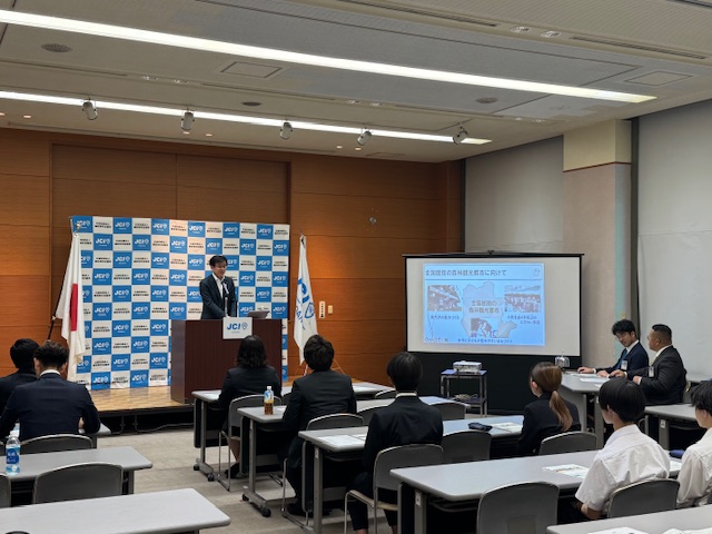 秦野青年会議所のバックパネルや、資料が投影されたスクリーンが設置された室内で、席に座ったスーツ姿の参加者が見ているなか、演台の前に立つスーツ姿の市長がマイクに向かって話をしている会議の様子の写真