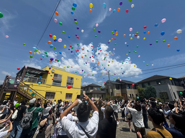 住宅街に囲まれたひまわり保育園の園庭で、快晴の空に向かって、たくさんの色とりどりの風船が放たれていく中、大勢の参加者たちがそれを見上げてスマートフォンをかざしている様子の写真