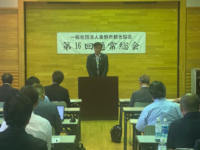 「一般社団法人秦野観光協会 第16回通常総会」と書かれた横断幕が貼られた木目の壁の室内で、演台に立つスーツ姿の市長が、着席した聴衆に向かって挨拶をしている写真