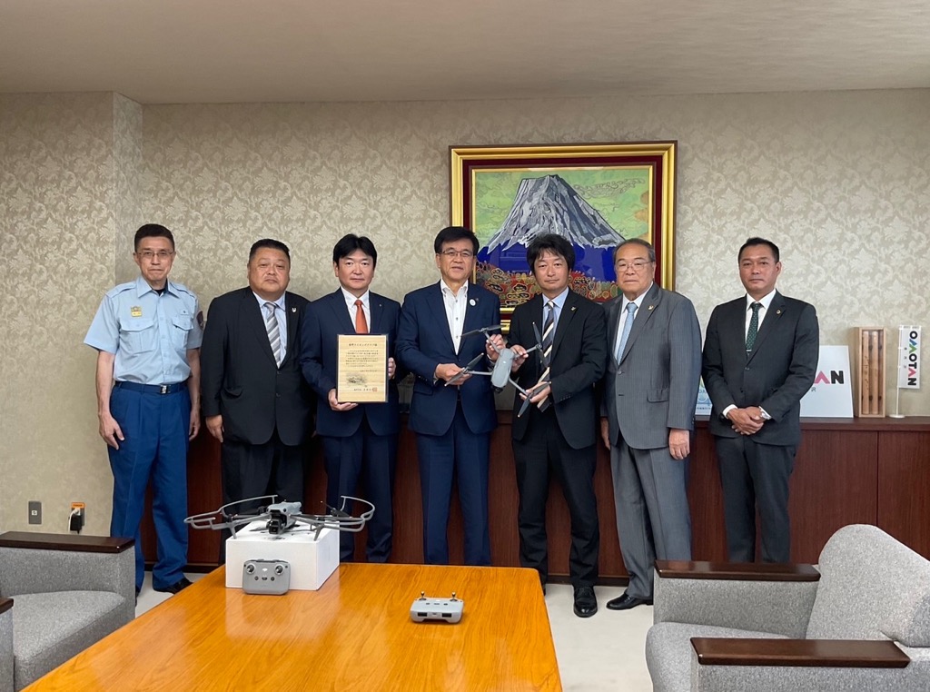 金色の額縁に入った山頂に雪が積もった大きな山の絵画を背景に、スーツ姿の6人の男性と制服姿の1人の男性が横一列に並び、中央の市長と代表の男性がドローンを持ち、左から3番目の男性が書類を持ち、手前のテーブルには寄贈されたドローンとリモコンが置かれている様子を写した記念写真