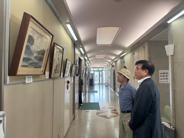 壁に絵画が飾られた細長い廊下で、帽子をかぶった男性とスーツ姿の市長の二人が、作品を鑑賞している様子を写した写真