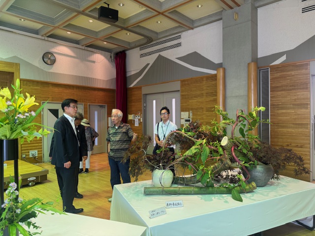 室内の展示会で、木の枝や葉を使った黒い壺や白い壺に生けられた作品、竹の上に生けられた生け花作品が展示された前で、スーツ姿の市長が男性から説明を受けながら鑑賞している華道展の様子を捉えた写真