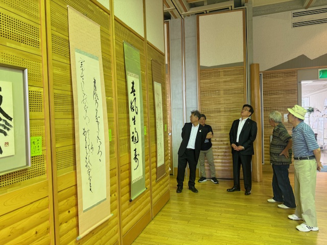 板をはめ込んだ壁に飾られた複数の掛け軸を、スーツ姿の市長を含む5人の来場者が鑑賞している書道展の写真