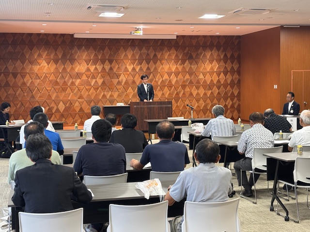 会議に参加している多くの聴衆を前に、三角形の模様が特徴的な壁を背景に演台に立つスーツ姿の市長が挨拶を行っている総会の様子の写真