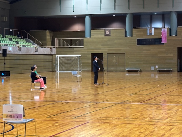 後方にはサッカーゴールと2階の観客席、そして話を聞く2人の関係者が椅子に座っている体育館で、フローリング床に立つスーツ姿の市長がマイクスタンドの前で挨拶を行っている様子の写真