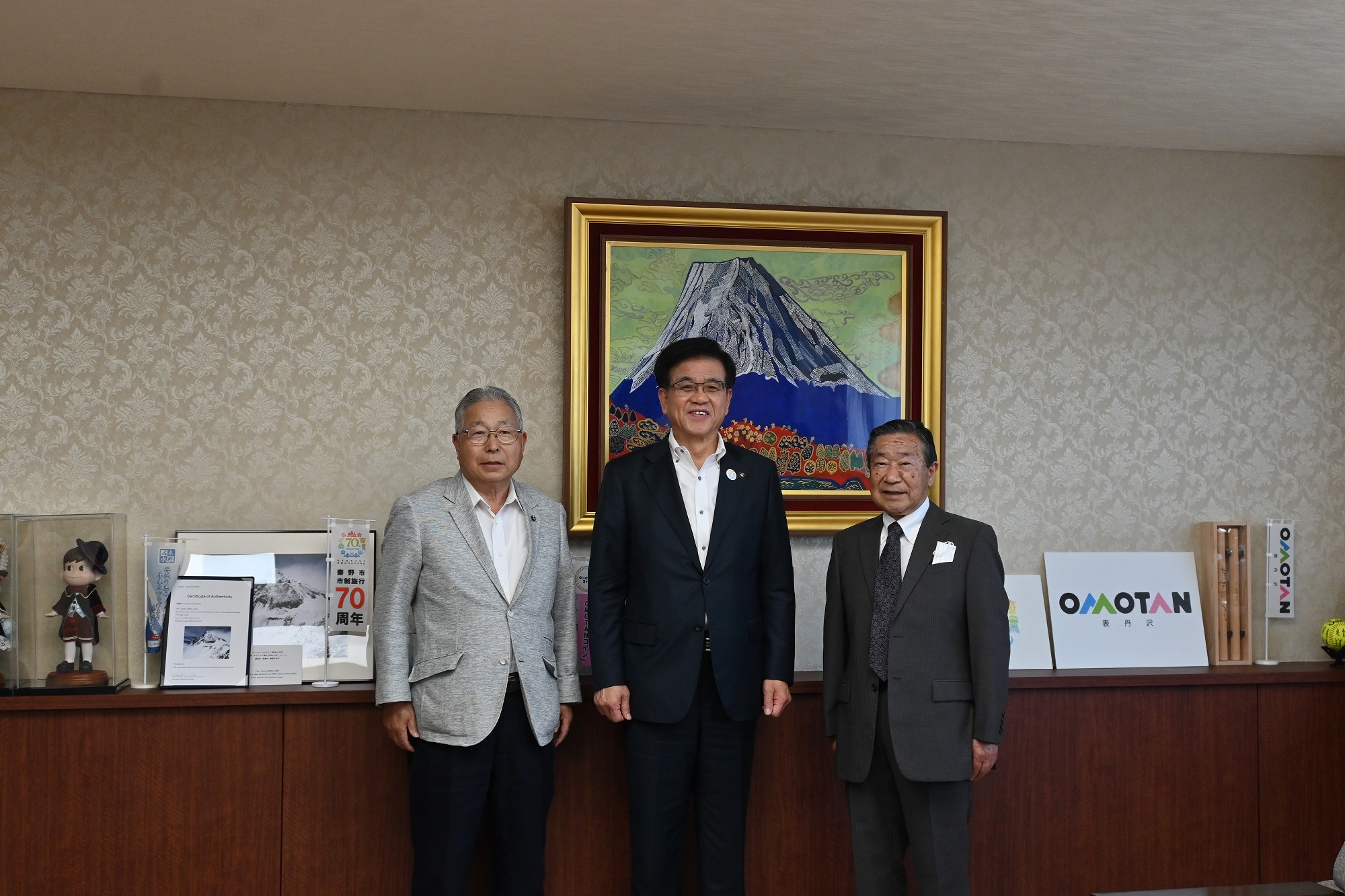 金色の額縁に入った山頂に雪が積もった大きな山の絵画が壁に飾られた落ち着いた背景の部屋で、スーツ姿の市長を中央に買い物クラブの2人の男性がカメラに向かって立ち、笑顔で記念撮影に応じている写真