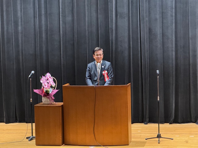 黒色の緞帳を背景にしたステージで、両脇に2台のマイクスタンドとピンクの胡蝶蘭が飾られた茶色の演台の前に立ち、笑顔で話す左胸に胸章リボンをつけたスーツ姿の市長の写真