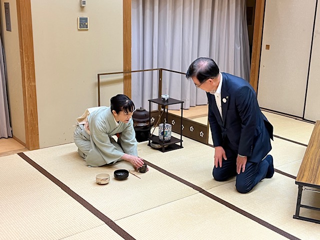 和室の畳の上で、薄い緑色の着物姿の女性がスーツ姿の市長に茶を点てており、茶道具を前に二人が向かい合って正座している様子の写真