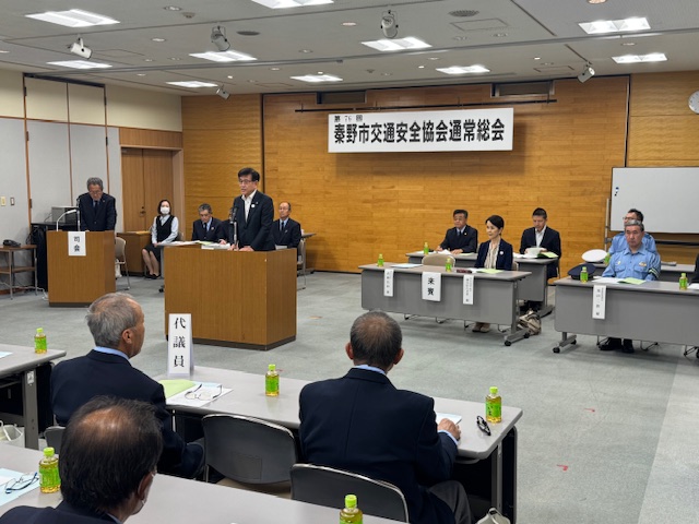 「第76回秦野市交通安全協会通常総会」と書かれた横断幕が掲げられた室内で、複数の関係者や参加者がテーブルを挟んで向かい合って座り、室内中央に設置された壇上の演台に立つスーツ姿の市長が挨拶を行っている様子の写真
