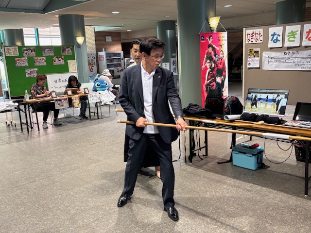 施設のロビーのような場所で、道着を着た男性指導者に見守られながら、スーツ姿の市長がなぎなたを構えているスポーツ体験イベントの様子の写真