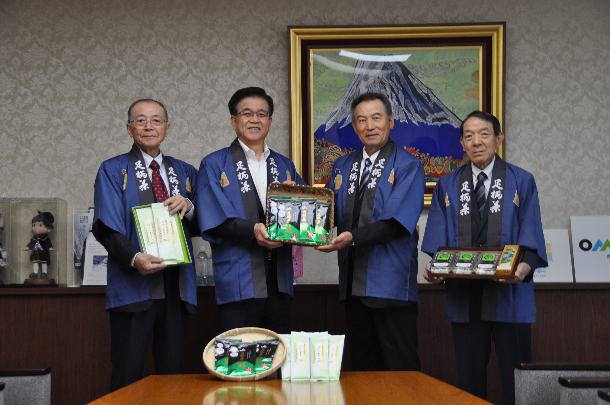 金色の額縁に入った山頂に雪が積もった大きな山の絵画が壁に飾られた落ち着いた背景の部屋で、「足柄茶」と書かれた紺色の法被を着た4人の男性が、中央のテーブルに置かれた2種類のお茶の商品と一緒にカメラに向かって立ち、中央に立つ市長と男性が黒と緑色のパッケージの商品が入ったカゴを一緒に持ち、左右に立つ2人の男性も他のお茶の商品を持って笑顔で写真に収まっている記念写真