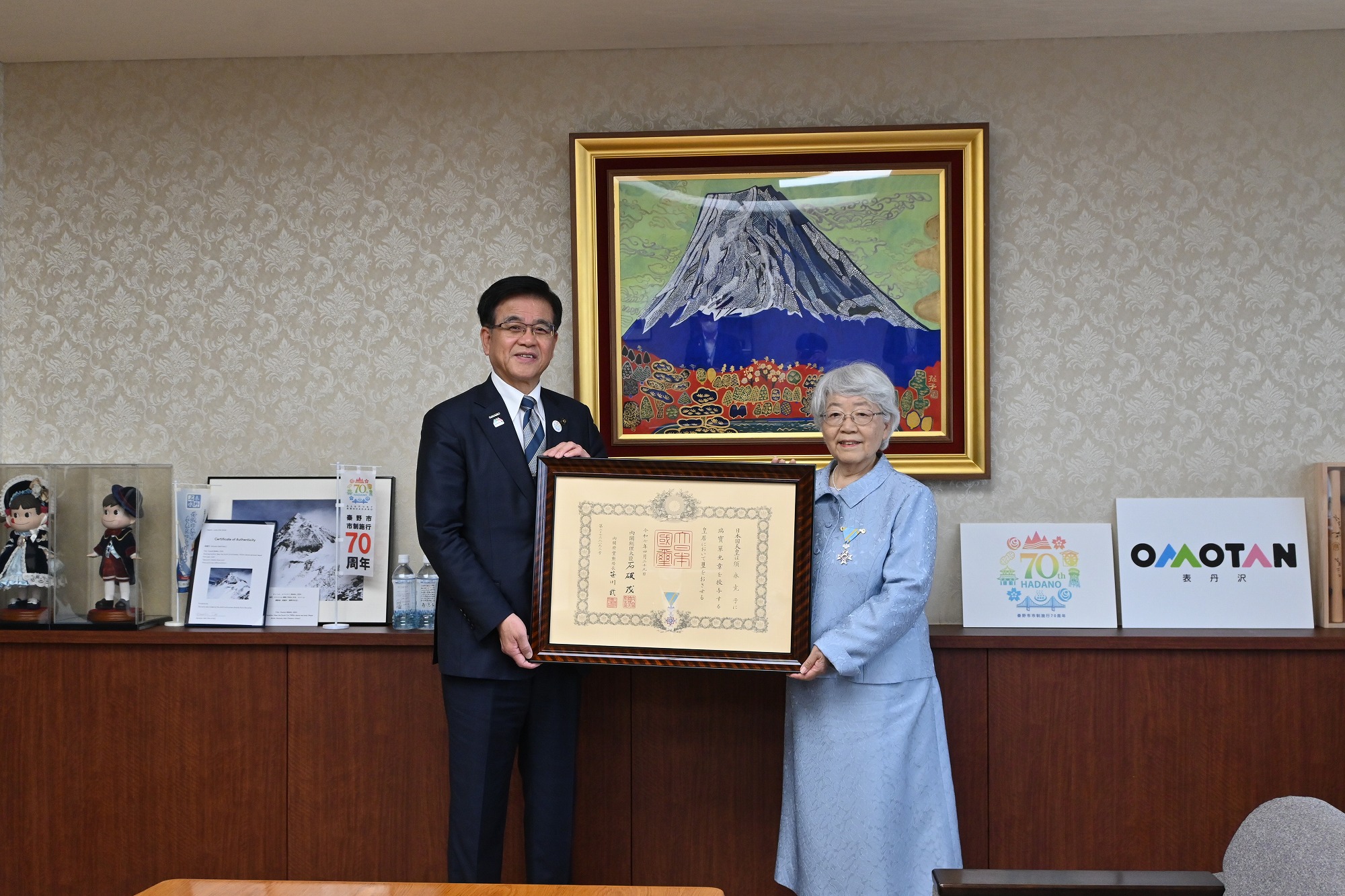 金色の額縁に入った山頂に雪が積もった大きな山の絵画が壁に飾られた落ち着いた背景の部屋で、スーツ姿の市長と、受賞した薄い水色のスーツを着た須永さんが、額縁に入った表彰状を一緒に持っている記念写真