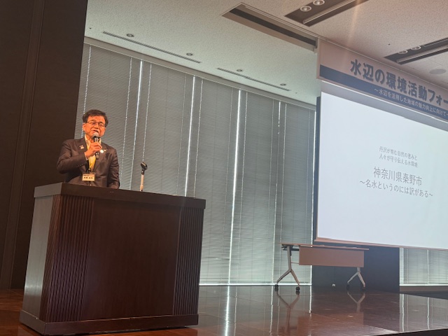背景には「水辺の環境活動フォーラム」のタイトルと「神奈川県秦野市」の文字が表示されたプロジェクタースクリーンが設置されたホールの演台で、スーツ姿の市長が話をしている写真