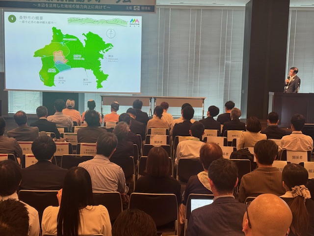 大勢の聴衆が集まった室内会場で、スクリーンに秦野市の地図が映し出され、壇上の演台に立つスーツ姿の市長がマイクを持って話している様子を後方から撮影した写真
