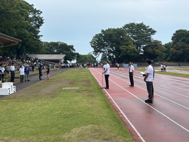 木々に囲まれた陸上競技場に大勢の参加者が立ち並び、赤色のトラックに3人の関係者とマイクスタンドの前に白いシャツを着た市長が立ち挨拶を行っている開会式の様子を捉えた写真