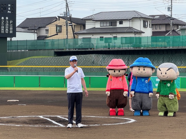住宅街に囲まれた野球場で、後ろに立つピンク色の帽子と服を着た女の子、青色の帽子と服を着た男の子、緑色の服を着た坊主頭の男の子の3体の可愛らしいマスコットキャラクターの着ぐるみが並んで見守るなか、中央にあるピッチャーマウンドで、白いシャツに野球帽をかぶった市長がマイクを持って挨拶を行っている様子の写真