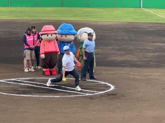 後ろに立つピンク色の帽子と服を着た女の子、青色の帽子と服を着た男の子、緑色の服を着た坊主頭の男の子の3体の可愛らしいマスコットキャラクターの着ぐるみと関係者の2人の女性が並んで見守り、傍で審判の男性が見ているなか、白いシャツに野球帽をかぶった市長がピッチャーマウンドからボールを投げている野球の始球式の様子の写真