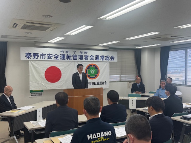 「令和7年度秦野市安全運転管理者会通常総会」と書かれた横断幕と2枚の旗が掲げられた室内で、多くの関係者が長机の席に座り見守るなか、前方に設置された演台に立つスーツ姿の市長が挨拶している総会の様子の写真