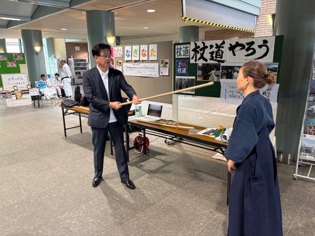 施設のロビーのような場所で、両手での丸木の杖を持ったスーツ姿の市長が、袴姿の女性から杖道の実演を教わっている様子の写真