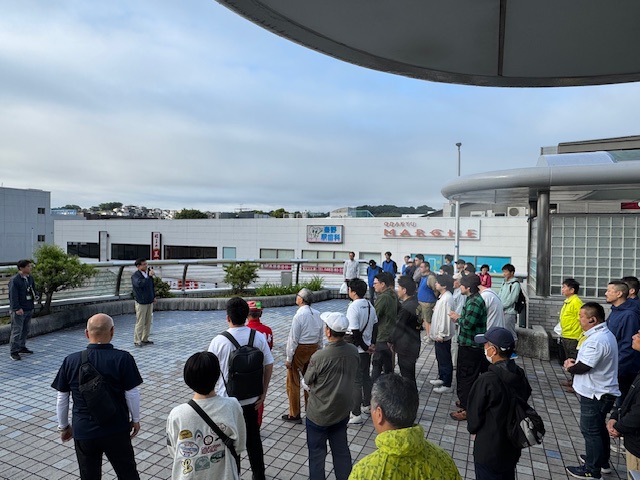 タイルが敷かれた屋外広場で、紺色のジャンパー姿の市長が、清掃活動に参加する大勢の参加者に向かってマイクを使い挨拶を行っている写真