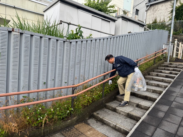 ゴミ袋を手に持った紺色のジャンパーを着た市長が、階段の手すり奥にある道端のゴミを腕を伸ばして拾っている様子の写真