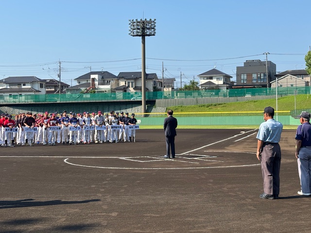 ナイターが設置された野球場で、スーツ姿の市長がマウンド近くに立ち、チームごとに整列した選手たちと審判員に向かって挨拶をしている、野球大会開会式の写真