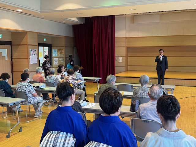 左側の入り口に立つ着物を着た女性、参加者や青色や白色の着物を着た人々が座って聞いている中、小さな舞台下に立つスーツ姿の市長が、マイクを持って挨拶を行っている様子の写真