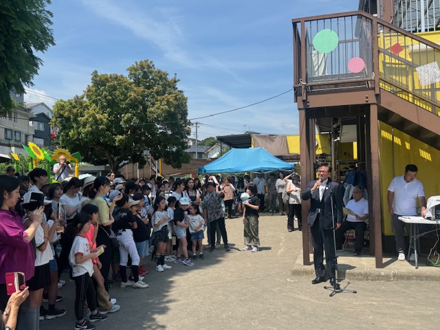 木々に囲まれた園庭で、木陰の下に集まった多くの親子連れや子どもたちが、茶色と黄色で施された園舎の前でスーツ姿の市長がマイクを持って挨拶しているのを熱心に聞いている記念式典の様子の写真