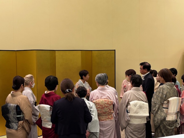 金屏風を背にした部屋で、スーツ姿の市長が複数の和服を着た女性たちに囲まれている親睦茶会の様子の写真
