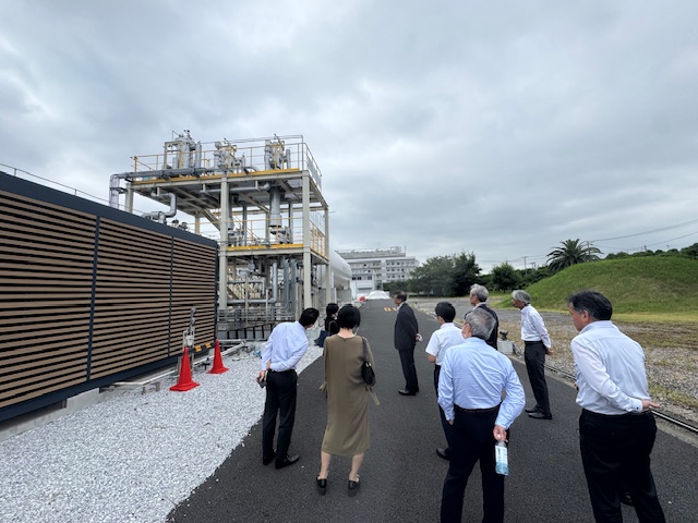 左側に黒と茶色の壁があり、右側に草が生えた小山がある広々とした敷地に建てられた工場で、大勢の男性と一人の女性が白とグレーのパイプが複雑に組み合わされた施設を見学している様子を捉えた写真
