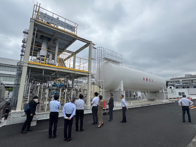 複数の男女が屋外に設置された「メタネーション設備」と書かれた2階建ての装置や、「水素ガス」と書かれた白い巨大なタンクを興味深げに視察している写真