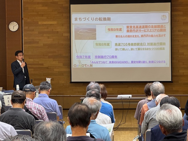 針が6時10分をさしている円形の時計が設置された壁際に、まちづくりの転換期についてのスライドがスクリーンに映し出され、左側の演台に立つスーツ姿の市長が、右手にマイクを持って聴衆に話している様子の写真