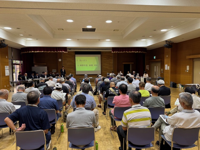 スクリーンに「あいさつ」と「秦野市長 高橋 昌和」の文字が映し出されている北地区の会場で、多くの人が椅子に座り、中央の壇上で話しているスーツ姿の市長を見ている様子を後方から撮影した写真