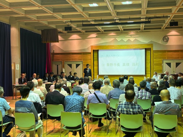 スクリーンに「あいさつ」と「秦野市長 高橋 昌和」の文字が映し出されている本町地区の会場で、大勢の参加者が室内に設置された椅子に座り、大きなスクリーンの左側に立つスーツ姿の市長がマイクを持って挨拶しているタウンミーティングの様子の写真