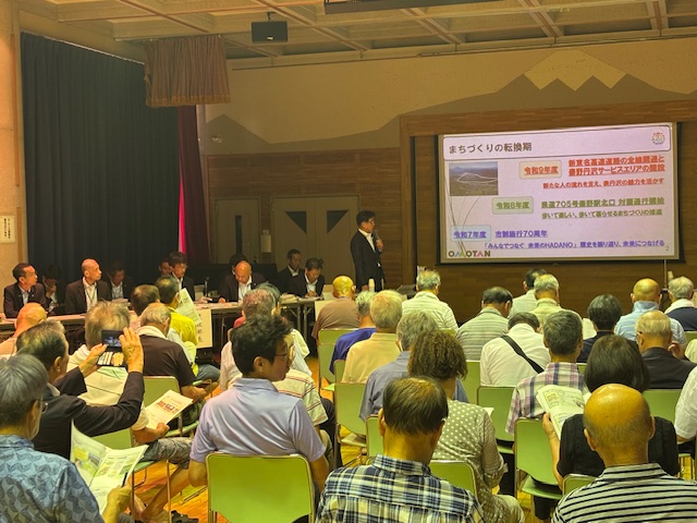 ホールの前方にプロジェクターを使い「まちづくりの転換期」というテーマの資料が映し出されたスクリーンの左側に立つスーツ姿の市長の話を、多くの聴衆が座って聞いているタウンミーティングの様子を後方から撮影した写真