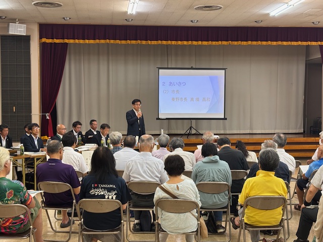 スクリーンに「あいさつ」と「秦野市長 高橋 昌和」の文字が映し出されている大根地区の会場で、小さな舞台の下に立つスーツ姿の市長がマイクで挨拶しており、多くの聴衆が着席して話を聞いているタウンミーティングの様子を後方から撮影した写真