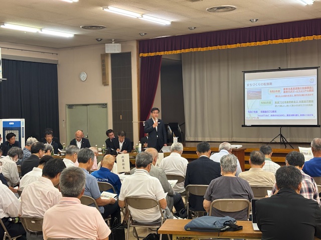 集会所で多くの聴衆が座る中、ステージ下に立つスーツ姿の市長がマイクを手に、プロジェクターに映し出された「まちづくりの転換期」というスライドについて説明している様子の写真