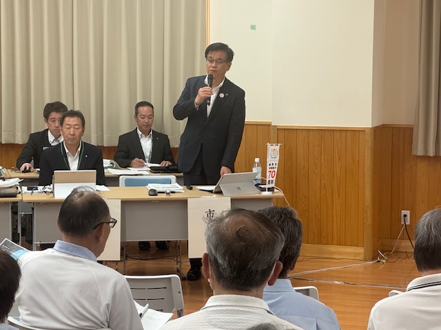 前列の机の席から立ち上がってマイクを持って話すスーツ姿の市長の話を、多くの聴衆が着席して聞いている、タウンミーティングの様子をアップで撮影した写真