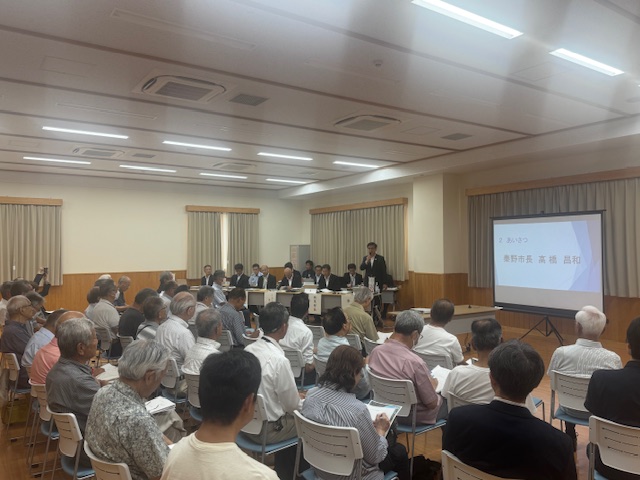 スクリーンに「あいさつ」と「秦野市長 高橋 昌和」と表示され、広間の前列の席から立ち上がったスーツ姿の市長が挨拶をしており、多くの人が座って話を聞いているタウンミーティングの様子を右後方から撮影した写真