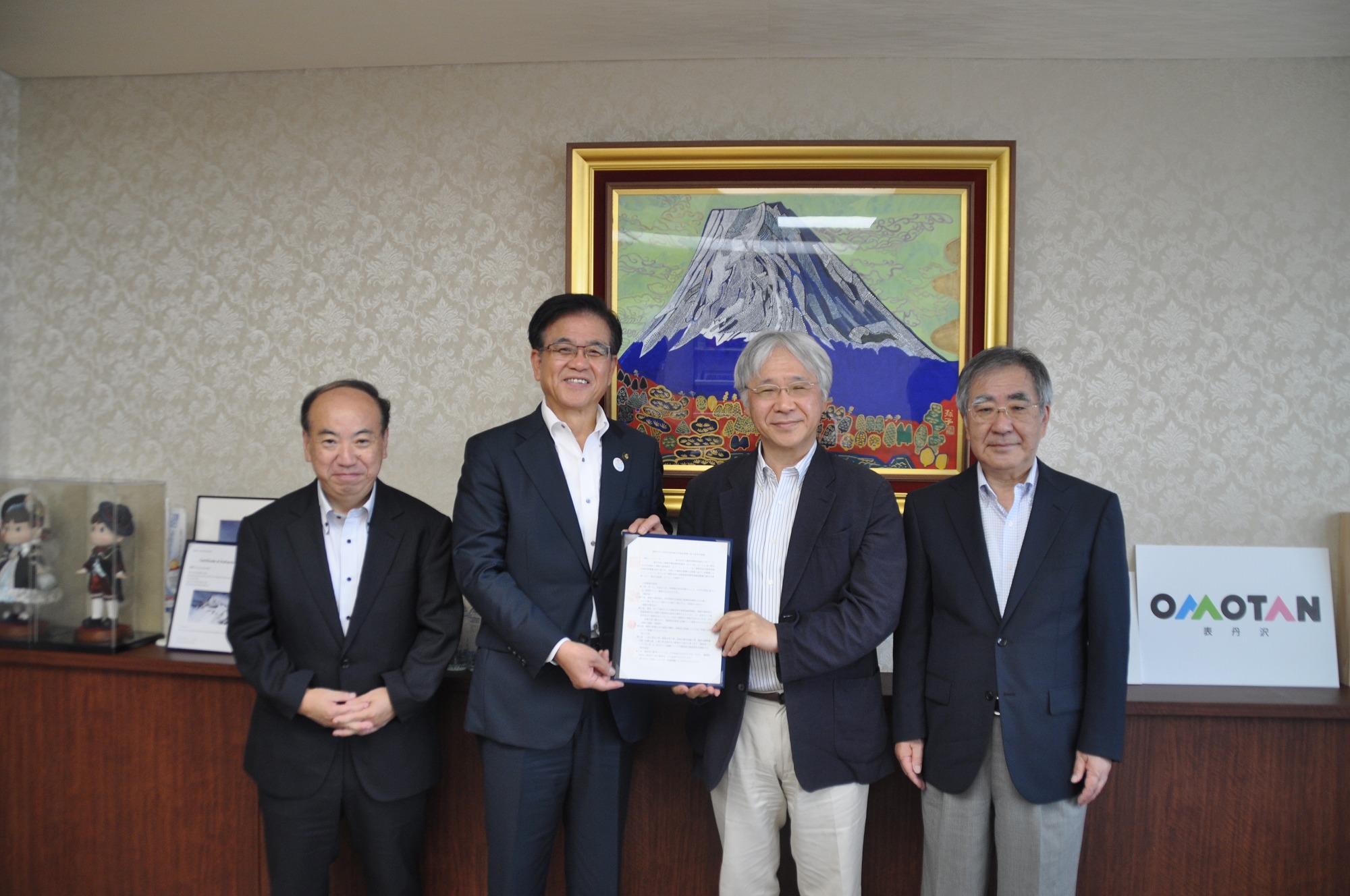 金色の額縁に入った山頂に雪が積もった大きな山の絵画が壁に飾られた落ち着いた背景の部屋で、スーツ姿の4人の男性が立ち、中央の市長と代表の男性2人が協定書のような書類を一緒に持ってカメラに向かい、笑顔で記念撮影をしている写真