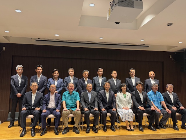 講堂の壇上で、複数人のスーツ姿の男性と女性が、前列の椅子に9人、後列に9人笑顔で並んでおり、集合写真を撮影している様子の写真